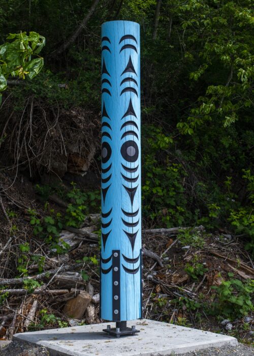 A blue totem pole with black tribal designs stands on a concrete base outdoors.