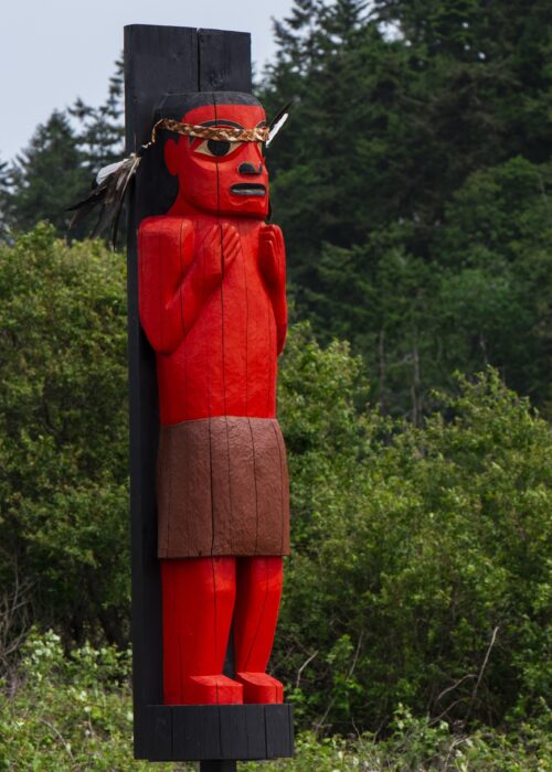 A carved red figure on a black post wears a brown skirt and a feathered headdress.