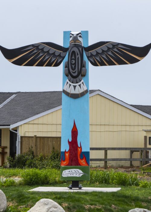 A colorful totem pole with an eagle at the top and flames at the bottom stands in a grassy area.