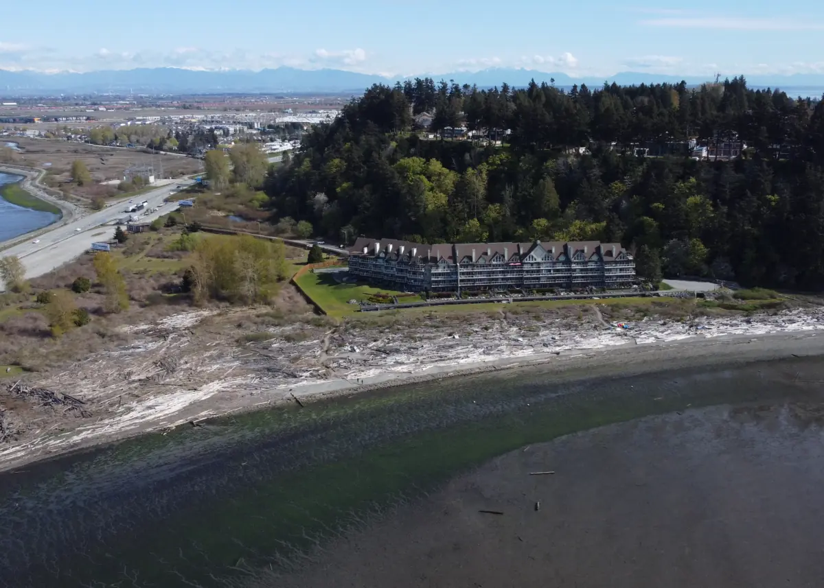 A long, multi-story building sits on a wooded bluff overlooking a beach covered in driftwood.