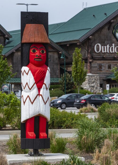 A red and white carved wooden totem pole stands in front of a large building with a green roof.