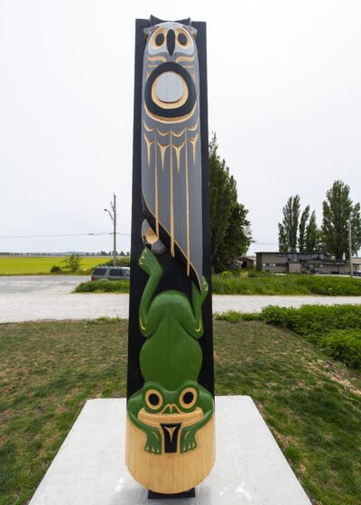 A totem pole carved with an owl at the top and a frog at the bottom, set against a rural landscape.